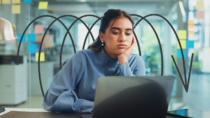 Employee resting her head on her hand while looking at a laptop, with hand-drawn downward arrows symbolising falling engagement.